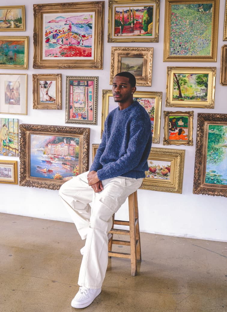 Camron De'Vine seated in front of a gallery wall of framed paintings